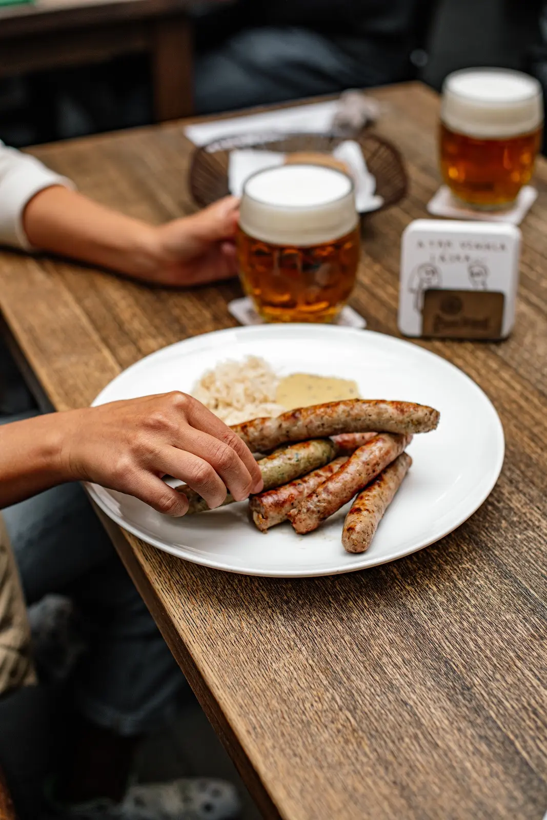 Grilované klobásy se zelím a hořčicí na talíři v restauraci Lokál, doplněné čepovaným pivem s bohatou pěnou.