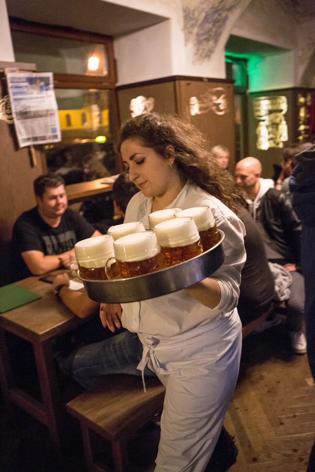 Číšnice v restauraci Lokál nese podnos s půllitry čerstvě načepovaného piva mezi hosty v zaplněném interiéru.