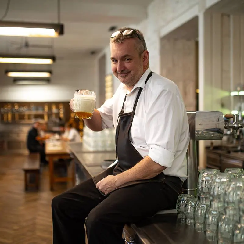 Smiling bartender in an apron sitting at the bar and holding a beer mug.